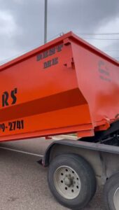 A Dump-IT Dumpster Rentals LLC orange dumpster being delivered and lowered from a truck in Laredo, TX.