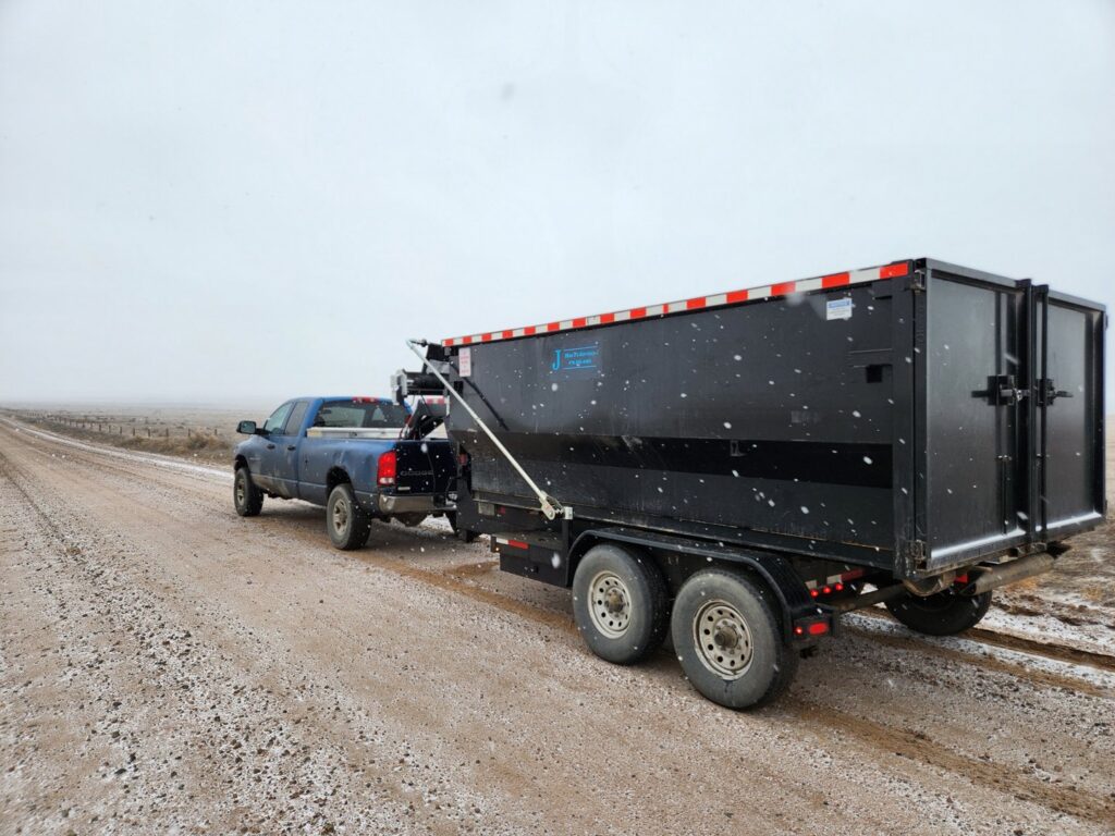 A Blue J's Services Dumpster Rental truck towing a large black dumpster on a snowy road in Greeley, CO, for delivery or pickup.