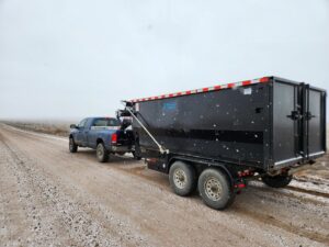 A Blue J's Services Dumpster Rental truck towing a large black dumpster on a snowy road in Greeley, CO, for delivery or pickup.