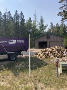 A McDonald Dumpsters purple dumpster being delivered near a wood pile and garage in Bar Nunn, WY.