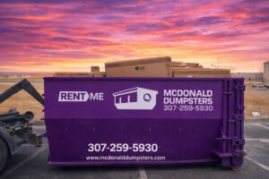 A McDonald Dumpsters purple dumpster filled with cardboard boxes under a sunset sky in Bar Nunn, WY.