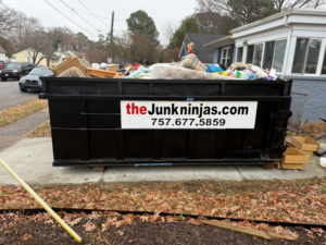 A black dumpster from The Junk Ninjas filled with various household junk items outside a home in Hampton Roads, VA.