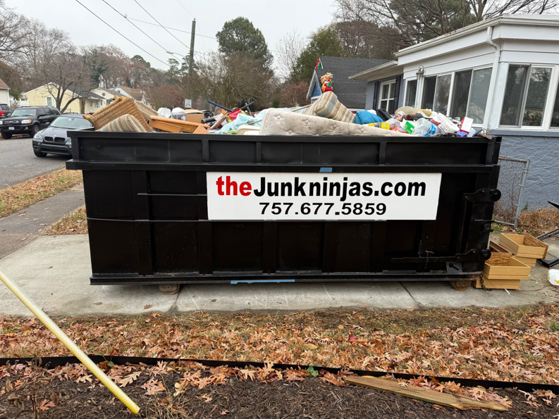 A black dumpster from The Junk Ninjas filled with various household junk items outside a home in Hampton Roads, VA.