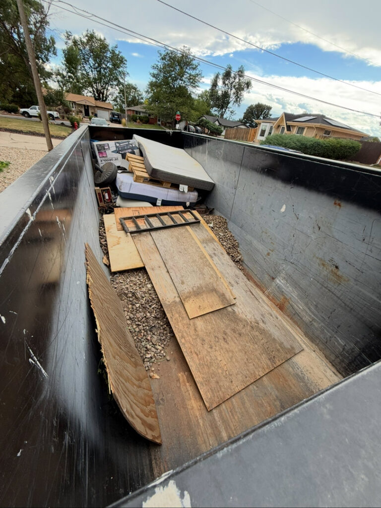 A Humpty Dump Roll-Offs & Dumpsters dumpster filled with various junk, debris, a mattress, and wood in Commerce City, CO.