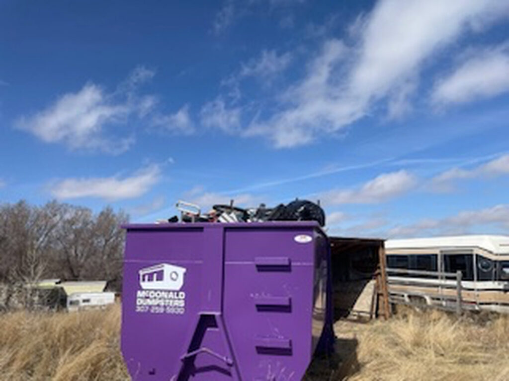 A McDonald Dumpsters purple dumpster filled with various junk and debris in a field in Bar Nunn, WY.