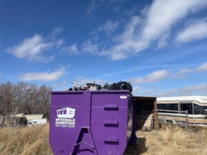 A McDonald Dumpsters purple dumpster filled with various junk and debris in a field in Bar Nunn, WY.