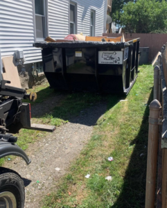 A black dumpster partially filled with junk, placed next to a house by Mini Dumpster Rentals in New Bedford, MA.