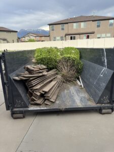 A black dumpster filled with yard waste and wood planks on a driveway by Utah Valley Dumpsters in Provo, UT.