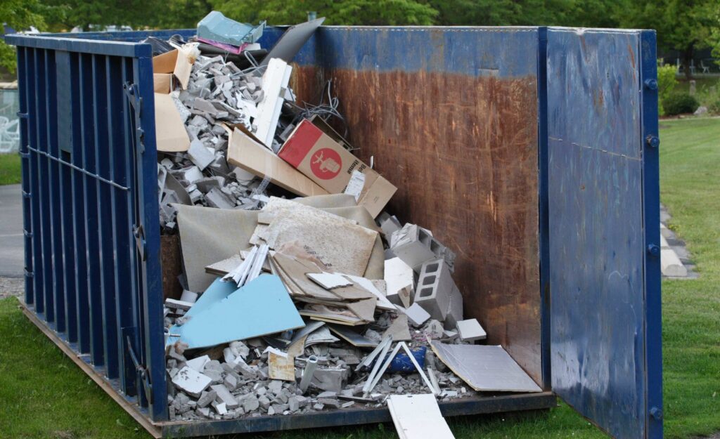 A blue roll-off dumpster filled with construction debris like concrete blocks, wood, and cardboard in a grassy area for junk removal in Sterling Heights, MI.