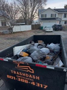 A TrashDash dumpster filled with household junk and debris in front of a residential home in West Haven, UT.