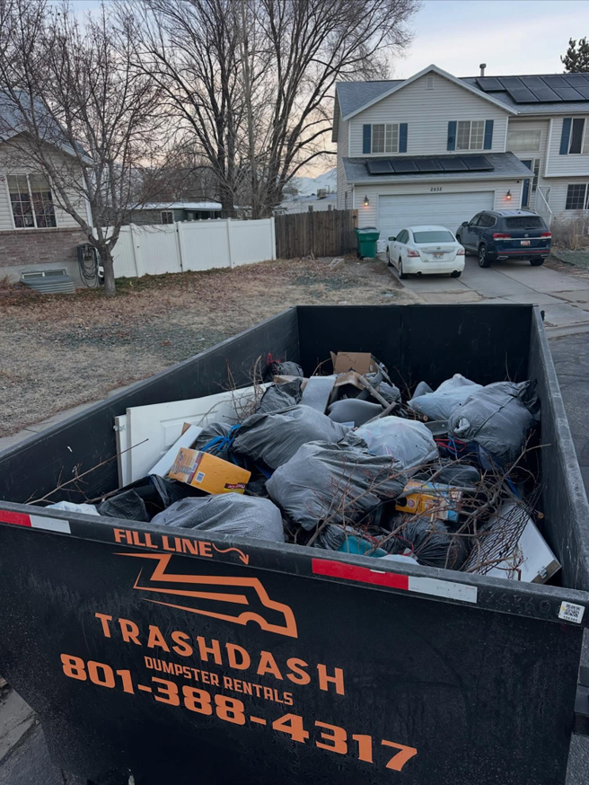 A TrashDash dumpster filled with household junk and debris in front of a residential home in West Haven, UT.
