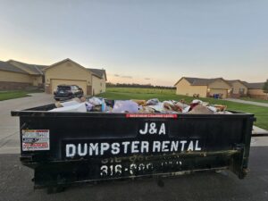 A J & A Haul Away & Dumpster Rentals black dumpster filled with household junk on a street in Derby, KS.