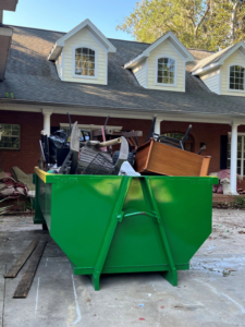 A green dumpster filled with various household junk and furniture in front of a residential home, provided by Wastecon in Harrisburg, PA.