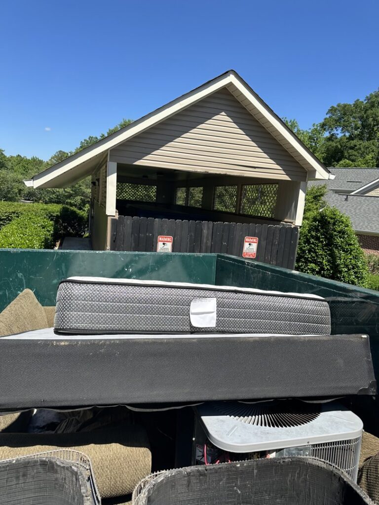 A green dumpster filled with a mattress, an AC unit, and other junk by Beetles Dumpsters Rental & Junk Removal in Huntsville, AL.
