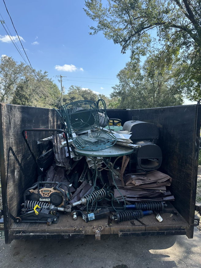 A dumpster full of miscellaneous junk including a patio chair, grill, tools, and car parts being removed by VMH services & hauling in Mulberry, FL.