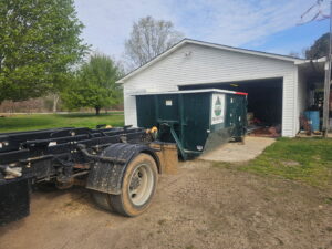 A green dumpster being placed or picked up from a garage by Beetles Dumpsters Rental & Junk Removal in Huntsville, AL.