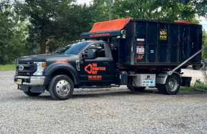 A branded junk removal truck from The Dumpster Guy, equipped with a roll-off container in Webster, MA.