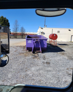 A purple All In Waste LLC dumpster placed in a gravel lot in Greenville, SC.