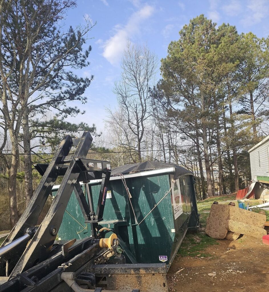 A green dumpster being loaded onto a truck, with another dumpster nearby, by Beetles Dumpsters Rental & Junk Removal in Huntsville, AL.