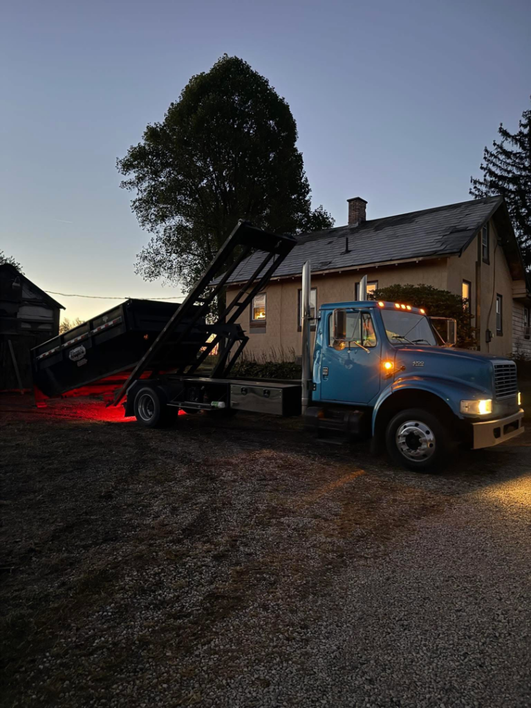 A blue roll-off truck loading or unloading a dumpster for junk removal services at dusk by Wagners Property Services LLC in Canton, OH.