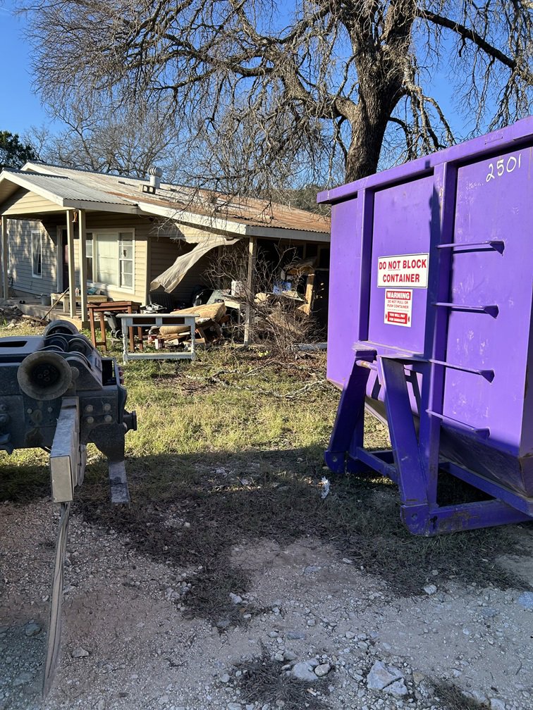 A purple dumpster from Liberty Hill Dumpster Rental next to a house with a large debris pile in Austin, TX