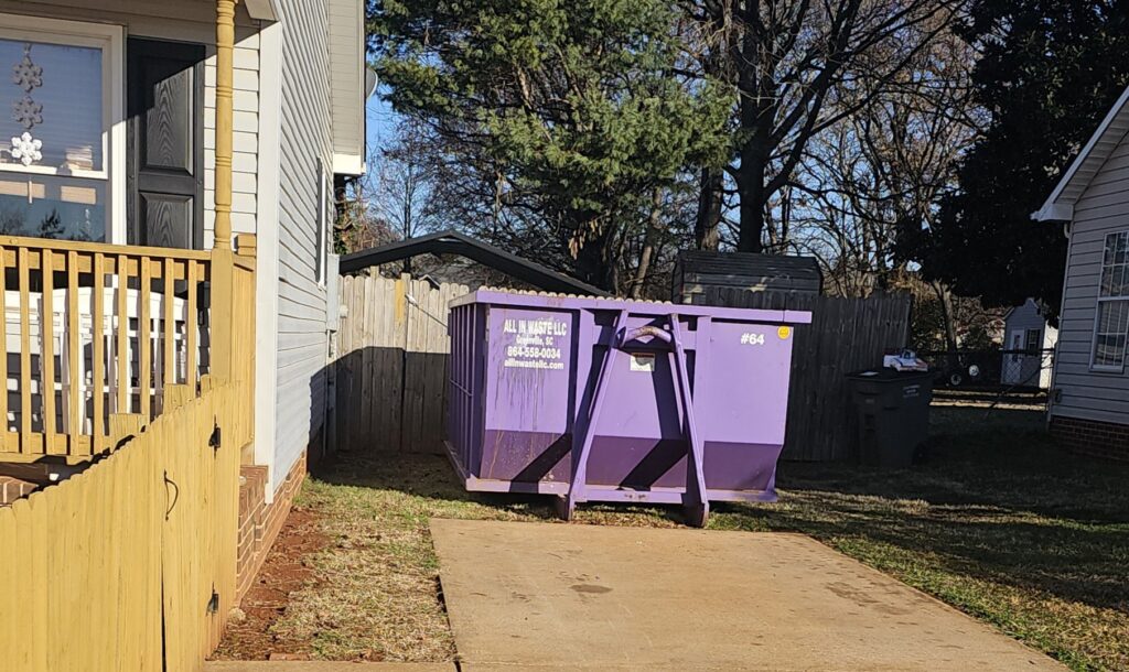 A purple All In Waste LLC dumpster placed next to a residential house in Greenville, SC.