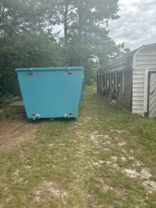 A blue dumpster placed next to an old shed for a cleanup project by Site Boss Dumpster Rentals in Raeford, NC.