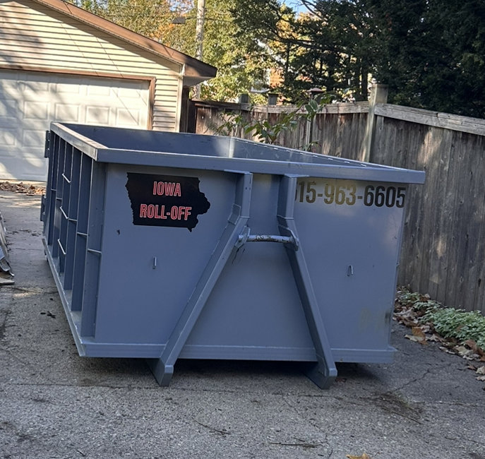 A grey roll-off dumpster placed on a paved driveway next to a wooden fence by Iowa Roll Off in Ankeny, IA.