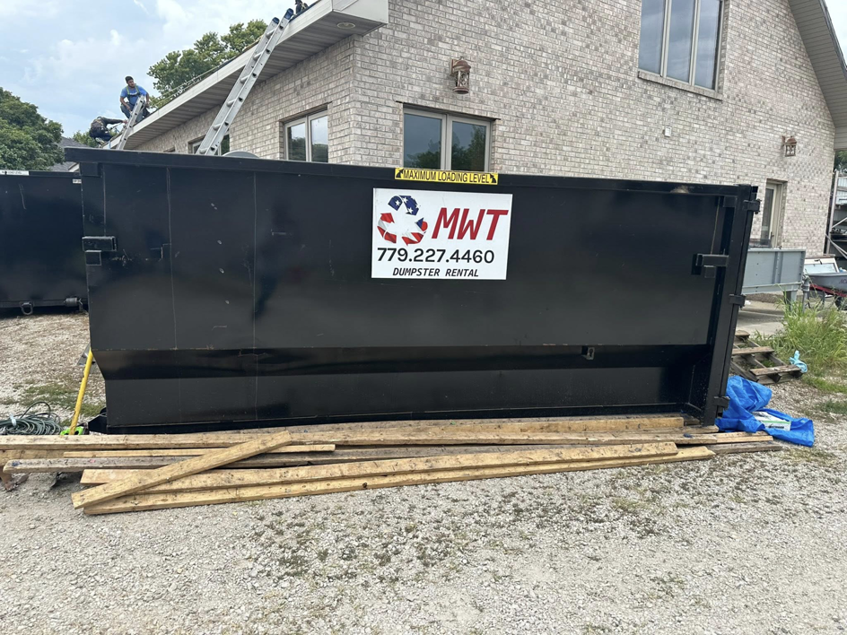 A large black MWT Dumpster and Hauling Services dumpster on site for junk removal at a residential property in Morris, IL.