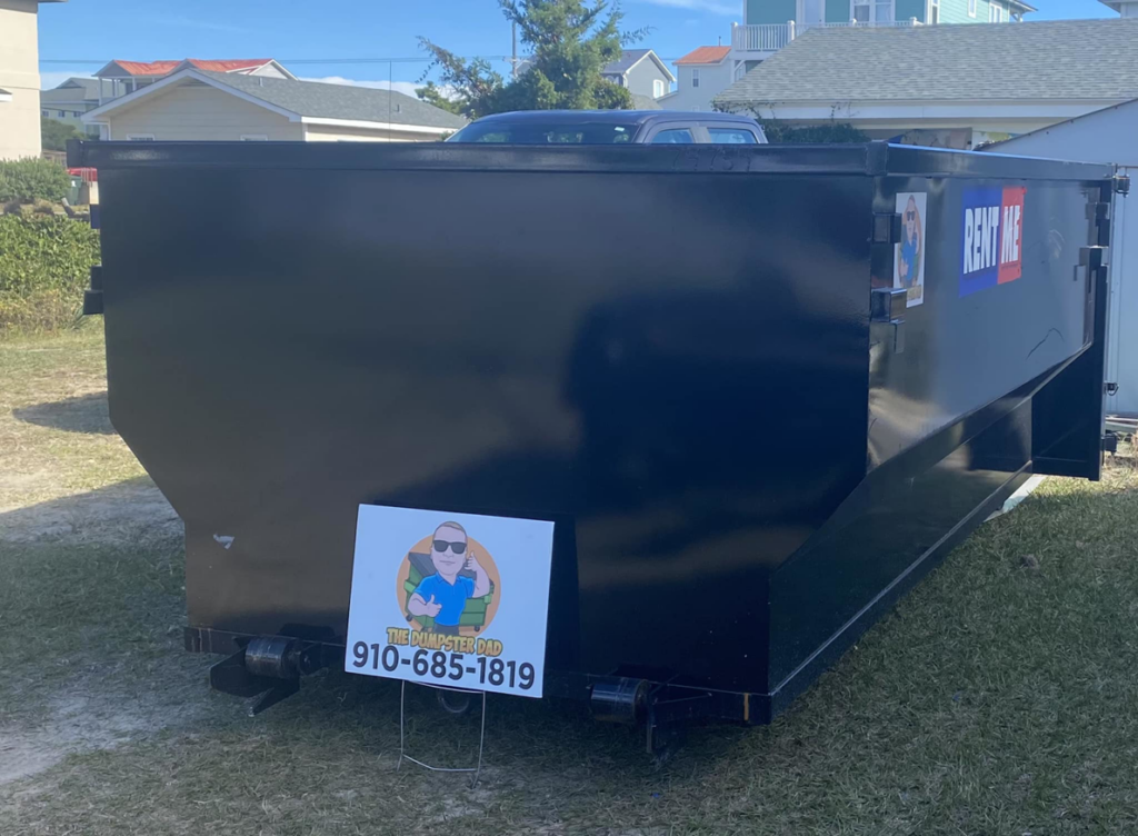 A black dumpster placed on a grassy area with a 'The Dumpster Dad' sign in front of it, in Wilmington, NC.