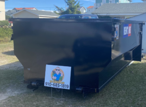 A black dumpster placed on a grassy area with a 'The Dumpster Dad' sign in front of it, in Wilmington, NC.