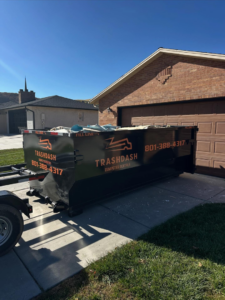 A TrashDash dumpster on a trailer parked in a residential driveway next to a garage in West Haven, UT.