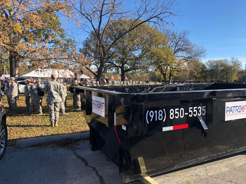 A black dumpster provided for waste management at an outdoor event by Patriot Dumpster Rental and Hauling in Tulsa, OK.
