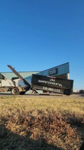 A J & A Haul Away & Dumpster Rentals truck picking up a black dumpster at a commercial location in Derby, KS.