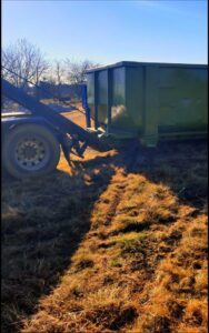 A FJD Waste Disposal and Recycling LLC truck picking up a green dumpster from a grassy area after a junk removal job in Dallas, TX.