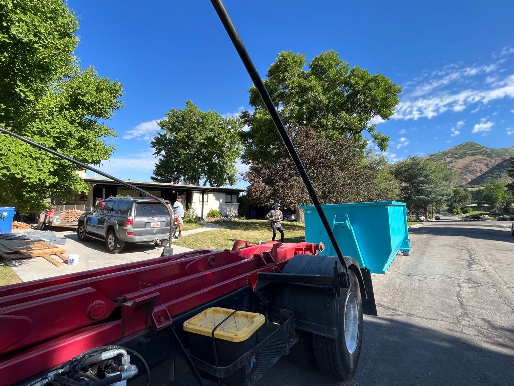A truck picking up a blue dumpster from a residential property, providing junk removal service by Blue Bin Dumpster Rentals in Salt Lake City, UT.