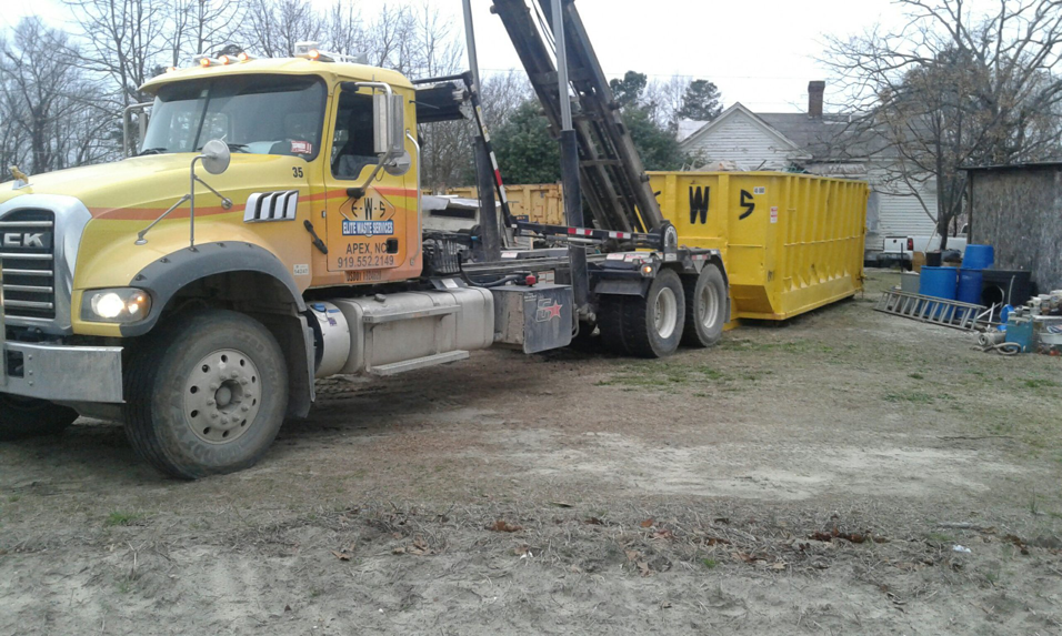 An Elite Waste Services truck picking up a yellow roll-off dumpster from a property in Raleigh, NC.