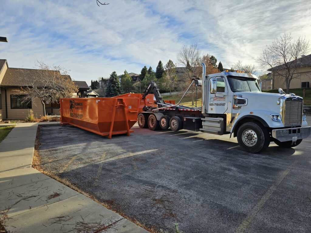 A Rushmore Dumpster truck placing an orange roll-off dumpster in a parking lot for a job in Rapid City, SD.
