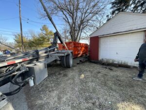 A truck preparing to place or pick up an orange dumpster next to a residential garage by Lil Man W/A Can LLC in Columbus, OH.