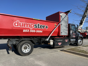 A red dumpster+ roll-off truck with a dumpster and portable toilet for junk removal in Columbia, SC.