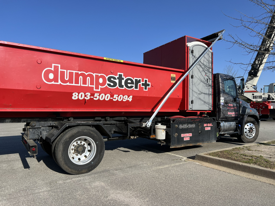 A red dumpster+ roll-off truck with a dumpster and portable toilet for junk removal in Columbia, SC.