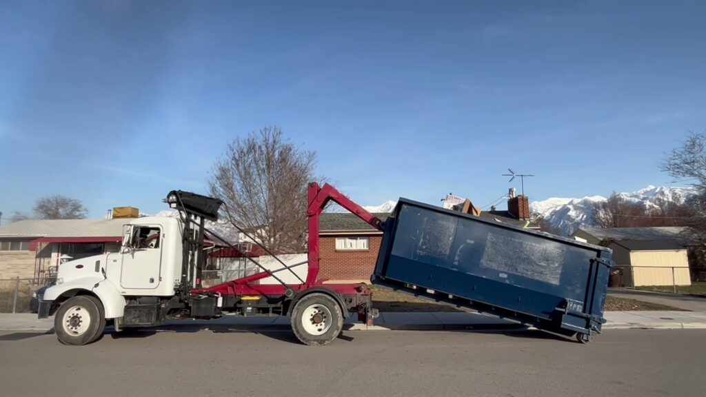 A truck removing a blue dumpster from a street, demonstrating the junk removal service of Blue Bin Dumpster Rentals in Salt Lake City, UT.