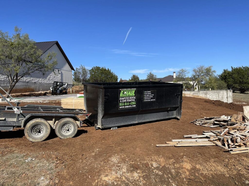 A G.I. HAUL Austin, TX branded dumpster on a trailer, positioned next to a pile of wood debris at a construction site.