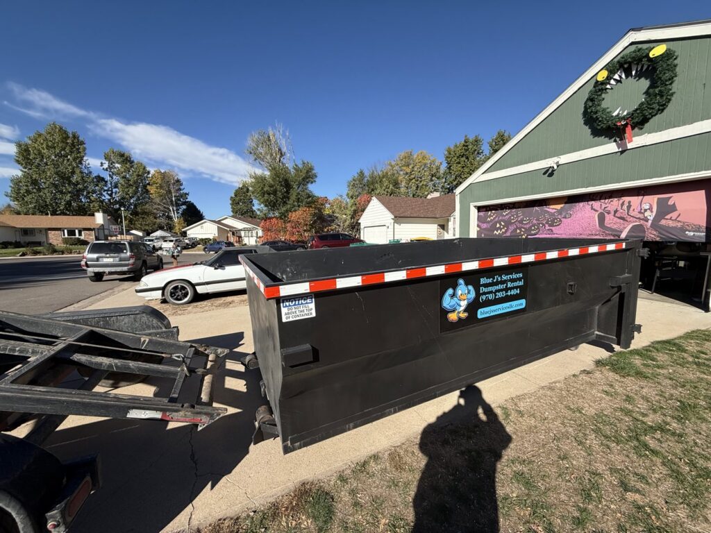 A Blue J's Services Dumpster Rental container parked in a residential driveway next to a garage in Greeley, CO.