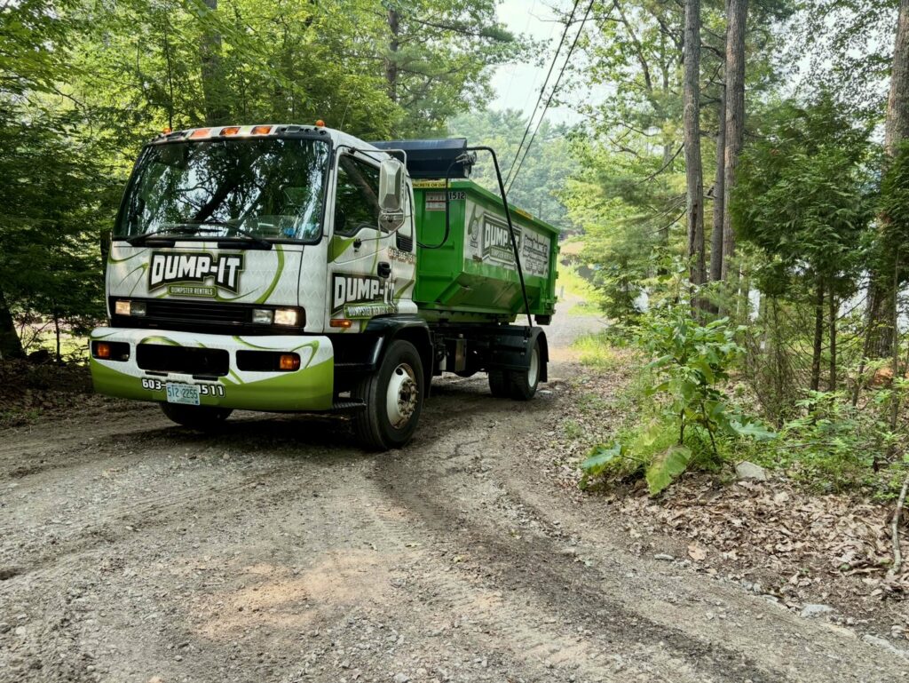 A Dump-It Dumpster Rentals truck with a green dumpster on its back, driving on a dirt road through a wooded area in Concord, NH.