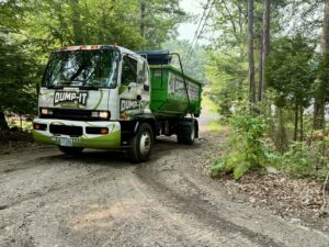 A Dump-It Dumpster Rentals truck with a green dumpster on its back, driving on a dirt road through a wooded area in Concord, NH.