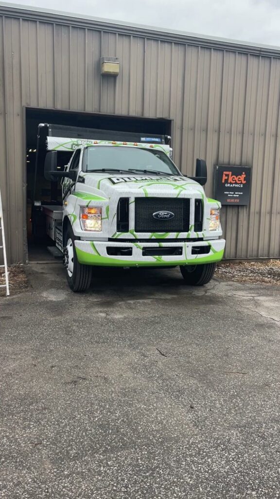 A Dump-It Dumpster Rentals truck, branded in green and white, exiting a garage in Concord, NH.