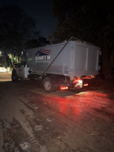 A Dump 'Em Dumpster Rentals truck with a dumpster parked on a dirt road at night in Charlotte, NC.