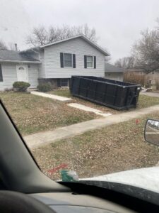 A black dumpster placed on a residential driveway for junk removal services by NID Dumpster Rental in Nashua, IA.