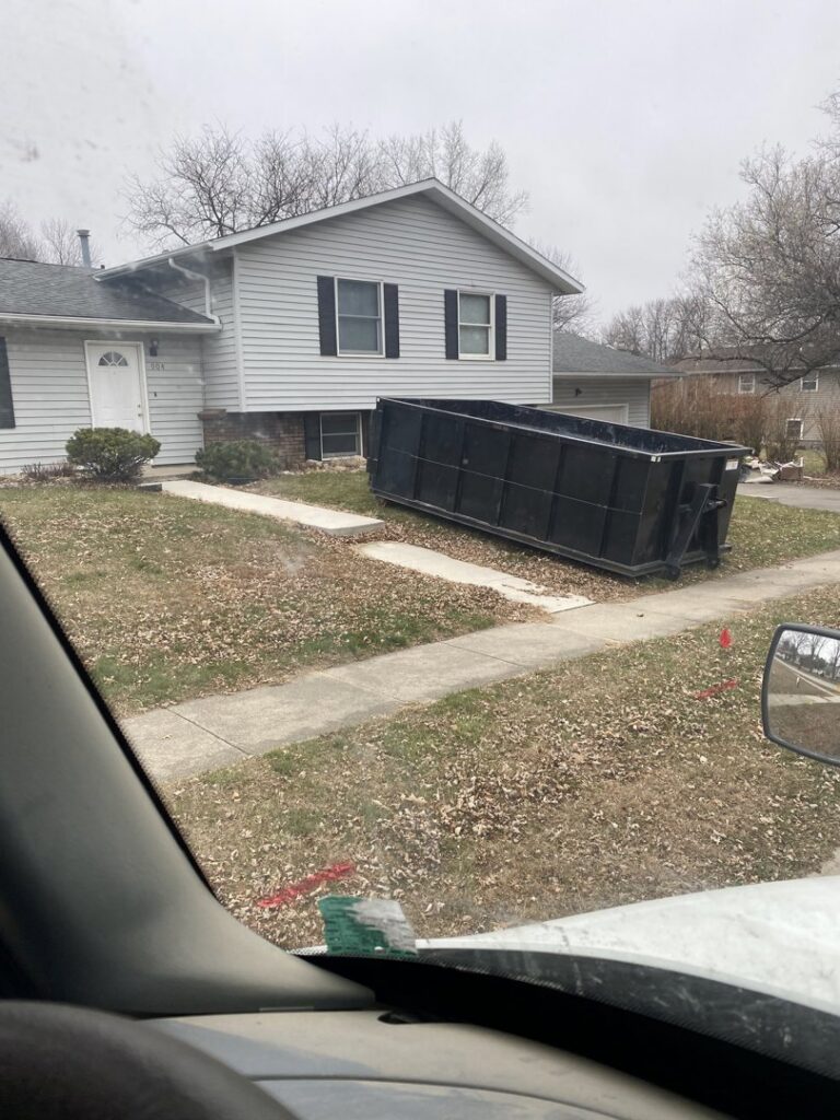 A black dumpster placed on a residential driveway for junk removal services by NID Dumpster Rental in Nashua, IA.